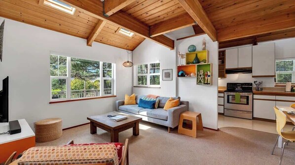 A modern living room with a sofa, coffee table, and a TV. There is a kitchen area with cabinets and a stove. Large windows provide natural light and views of the outdoors.