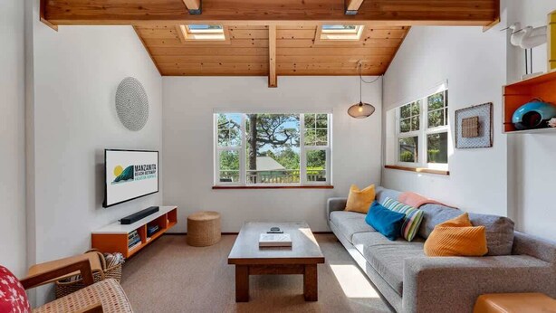 Ein modernes Wohnzimmer mit Holzboden, einem grauen Sofa mit bunten Kissen, einem Flachbildfernseher, einem Couchtisch und einem großen Fenster mit Blick auf Bäume.
