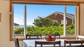 A dining area with a table and chairs, a bowl of fruit, and a large window with a view of a house and greenery. - Forever Manzanita by Manzanita Beach Getaway: Ocean View, Dog-Friendly, Hot Tub (Manzanita)