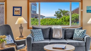 A living room with a grey sofa, patterned cushions, a lamp, a coffee table with a book, and a large window with a view of greenery and a building. - Forever Manzanita by Manzanita Beach Getaway: Ocean View, Dog-Friendly, Hot Tub (Manzanita)