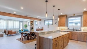 A spacious kitchen with wooden cabinets, a central island, pendant lighting, and a view to the living area.