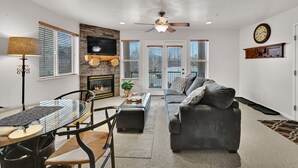 A modern living room with a fireplace, a ceiling fan, and a large window. - MH603 | Pinecone Retreat: Adventure Awaits in Eden, UT! (Eden)