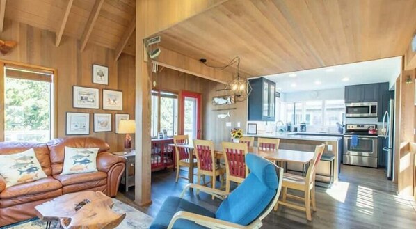 L’intérieur chaleureux d’un chalet avec un plafond en bois, un coin repas avec des chaises rouges, une cuisine équipée d’appareils modernes et un salon avec un canapé en cuir et des tableaux encadrés.