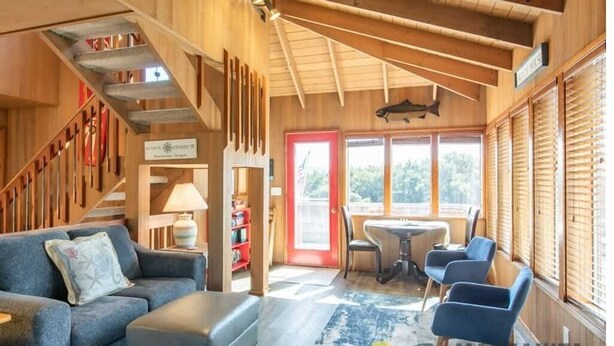Ein gemütliches Zimmer mit einer Couch, einem Hocker und einem kleinen Tisch mit Stühlen. Im Hintergrund ist eine Treppe mit Bücherregalen zu sehen.
