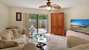 A living room with a glass-top coffee table, beige sofa, wooden cabinet, ceiling fan, and a TV on a stand.