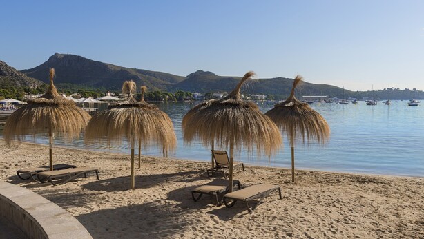 A beach with thatched umbrellas, lounge chairs, and a calm sea with boats. - Villa Can Oronella by Balearic Villas (Pollensa)