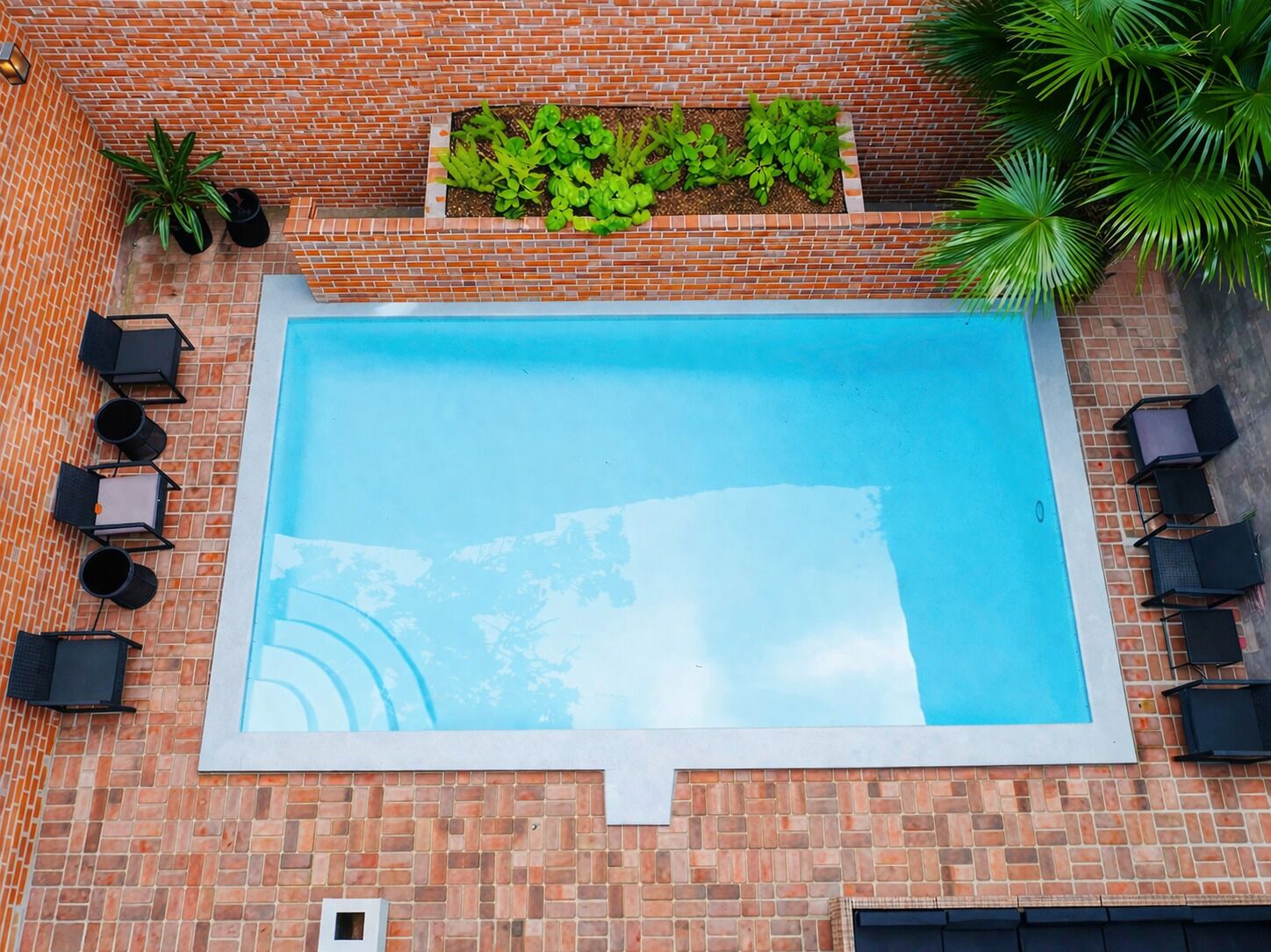 Una piscina rectangular rodeada por un muro de ladrillo, un macetero con plantas verdes y sillones de descanso negros alrededor de la piscina.