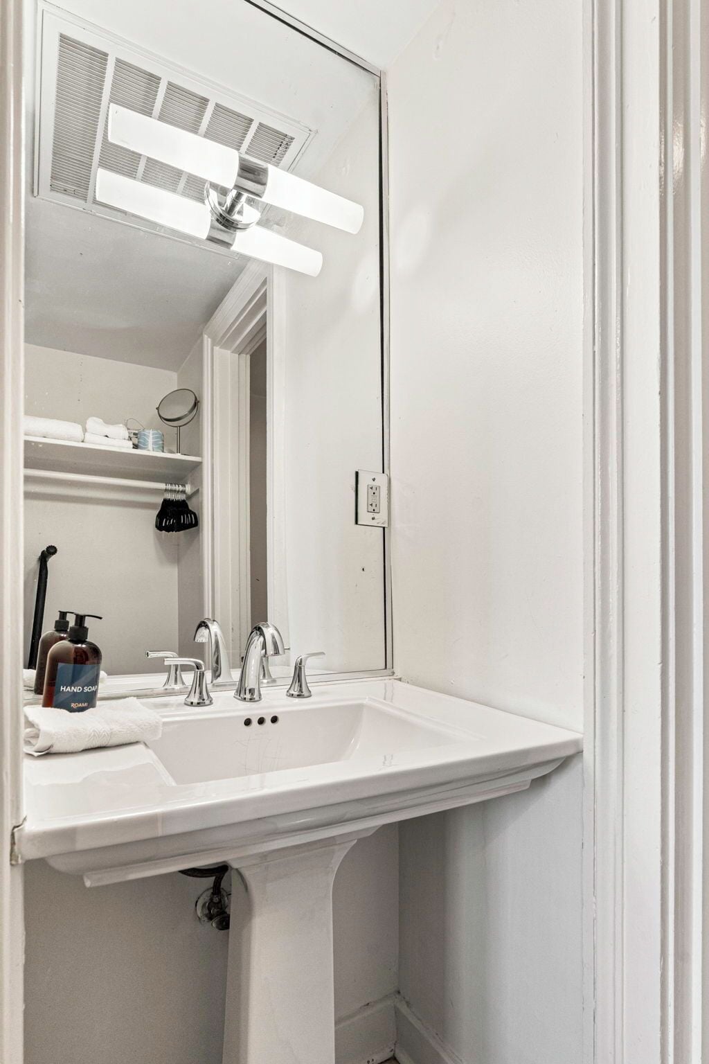Une salle de bain avec un lavabo blanc, deux robinets, un miroir et une grille d’aération au-dessus du lavabo.