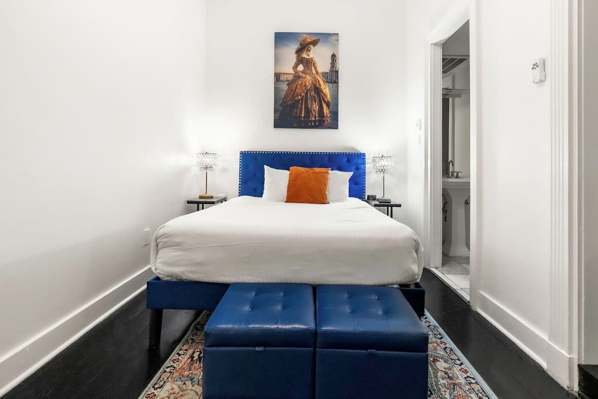 A bedroom with a blue headboard, white bedding, a bench, a painting, and a patterned rug.