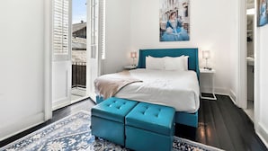 A bedroom with a bed, a blue bench, a painting, and a view of the outside. - Roami at St. Helene (New Orleans)