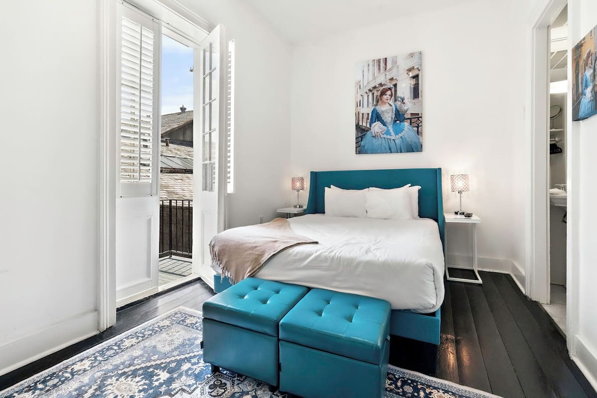 A bedroom with a bed, a blue bench, a painting, and a view of the outside.