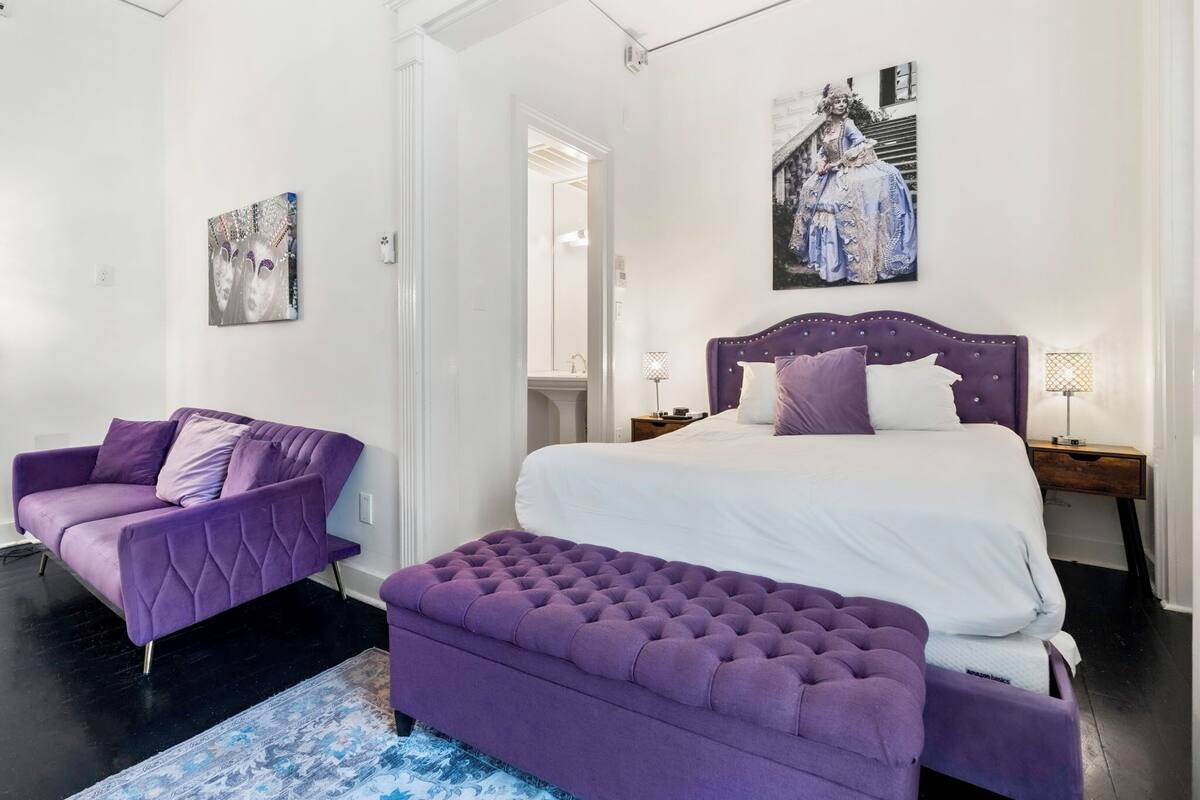 A bedroom with a bed, a purple sofa, a matching ottoman, and a painting on the wall.
