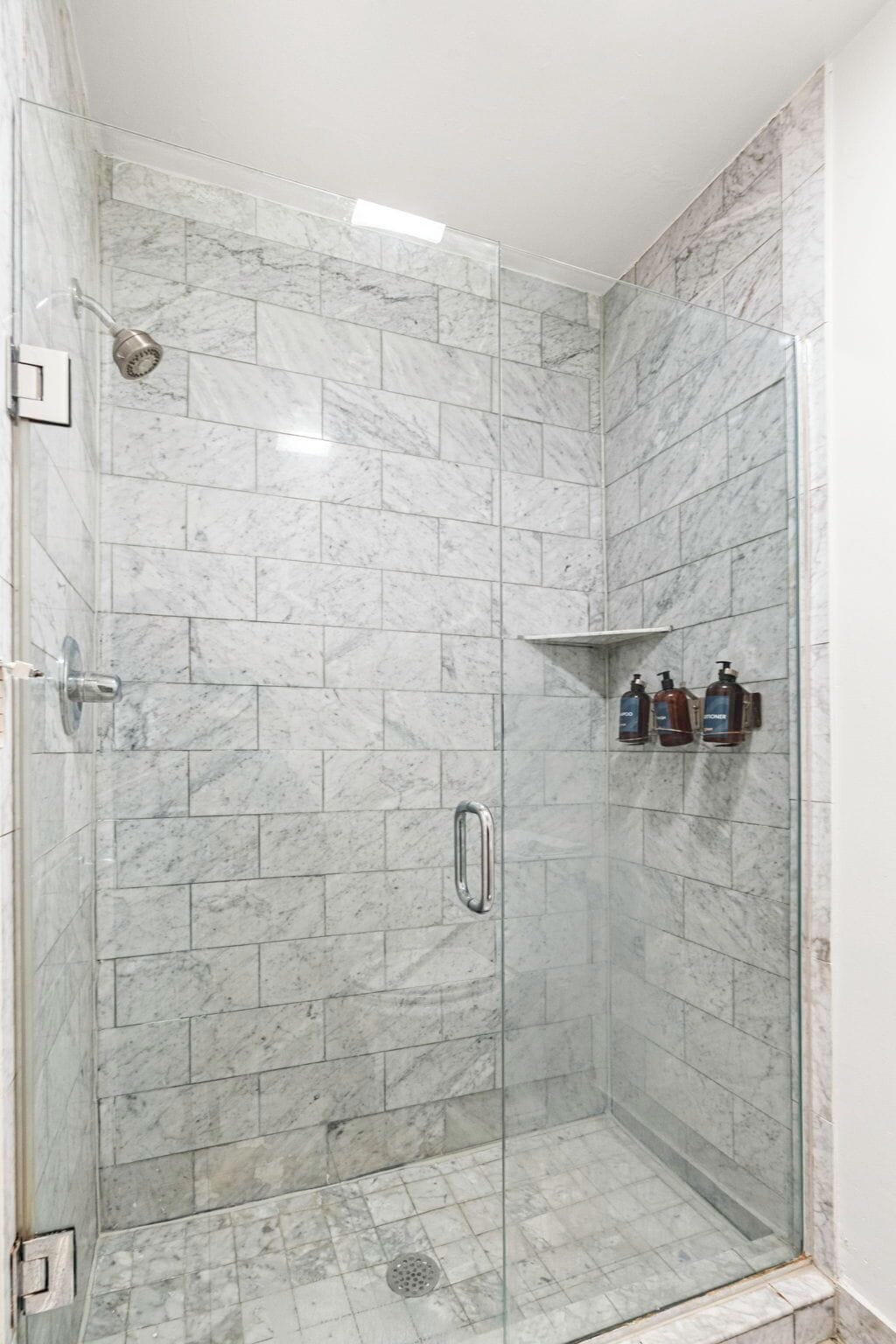 A glass-enclosed shower stall with marble tiles, a showerhead, a handheld shower, and two bottles of lotion on a shelf.