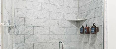A glass-enclosed shower stall with marble tiles, a showerhead, a handheld shower, and two bottles of lotion on a shelf.