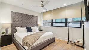 A modern bedroom with a large bed, a ceiling fan, a window with a view, and a nightstand with a lamp. - The Eleanor 4BD/4B - All King Bedrooms, City View (New Orleans)