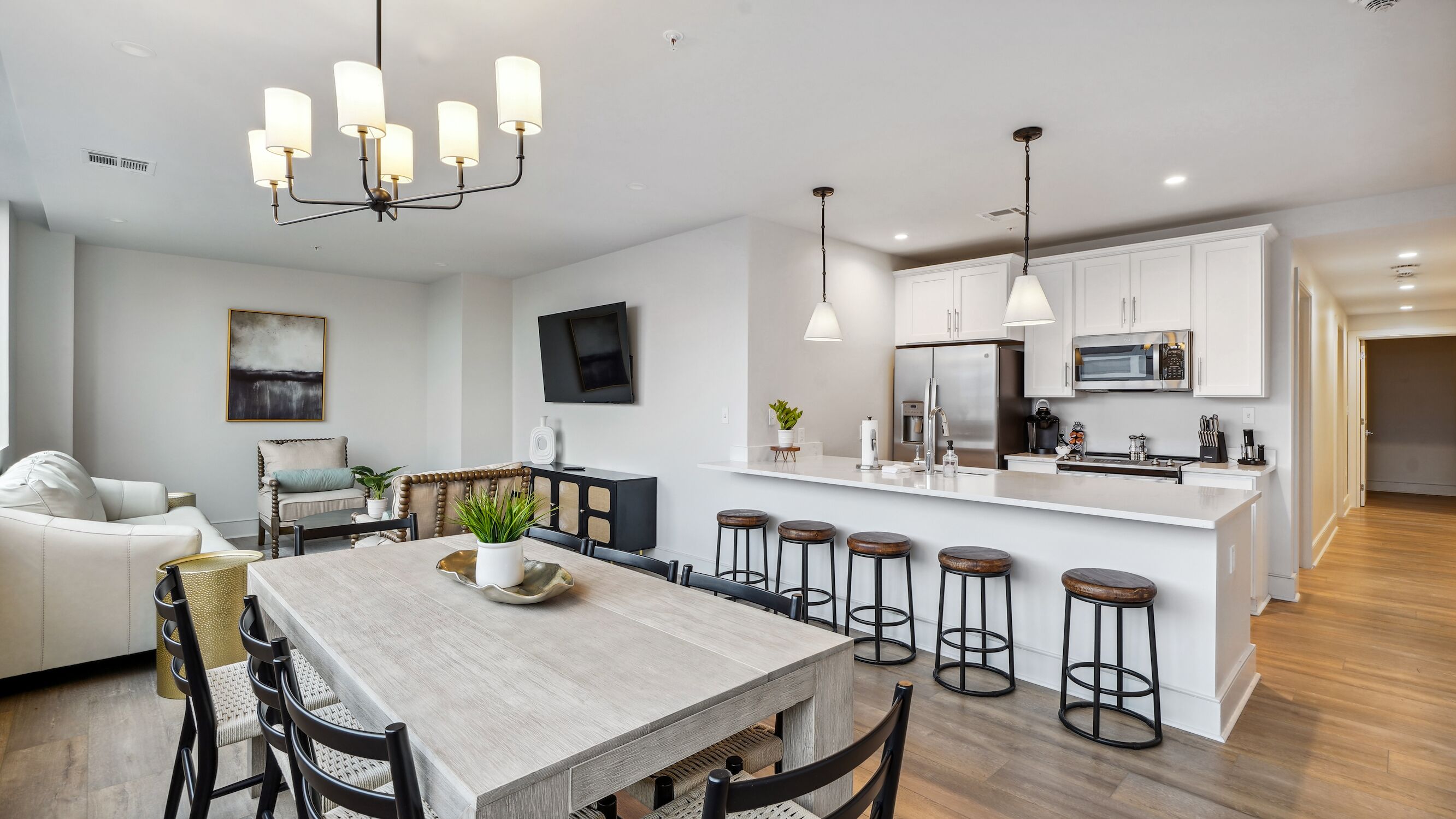 A modern kitchen with white cabinets, a central island with stools, a dining area with a wooden table, and a living room with a sofa and a TV.