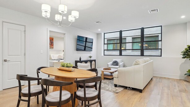 A modern living room with a round wooden table, four chairs, a white sofa, and a large window with a view of a brick building.