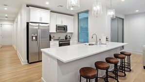 Een moderne keuken met witte kastjes, roestvrijstalen apparatuur, een groot eiland met krukken en hanglampen.