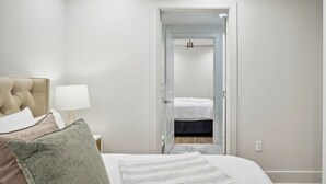 A bedroom with a bed, a nightstand, a lamp, and a door leading to another room. - The Eleanor 5BD/3B - 4 Kings & 2 Queens (New Orleans)