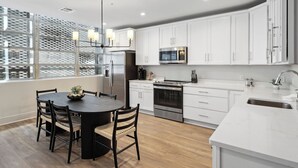 A modern kitchen with white cabinets, stainless steel appliances, a round dining table, and a large window with a patterned view. - The Eleanor 5BD/3B - 4 Kings & 2 Queens (New Orleans)