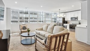 A modern living room with a beige sofa, a round coffee table, and a wicker chair. A kitchen with white cabinets and stainless steel appliances is visible in the background. - The Eleanor 5BD/3B - 4 Kings & 2 Queens (New Orleans)