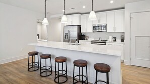 A modern kitchen with white cabinetry, a stainless steel refrigerator, a large island with stools, and pendant lighting. - The Eleanor 6BD/4B - Sleeps 16 (New Orleans)