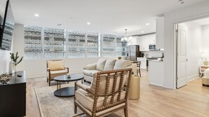 A modern living room with a sofa, armchair, and coffee table, a kitchen in the background, and large windows with a patterned view.