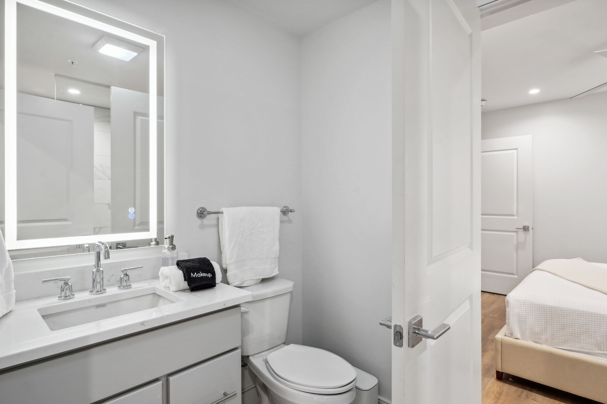 Un baño moderno con lavamanos, espejo y retrete blancos. Se ve una cama en la habitación contigua.