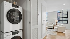 A modern laundry room with stacked washing machines, a closed door, and a living area with a sofa and a lamp. - The Eleanor 5BD/3B - 4 King Bedrooms & F/F Bunkbed (New Orleans)