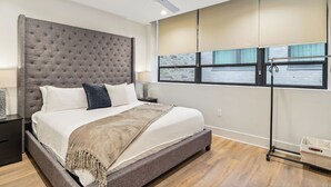 A modern bedroom with a tufted headboard, a large window with blinds, a ceiling fan, and a bedside table with a lamp.