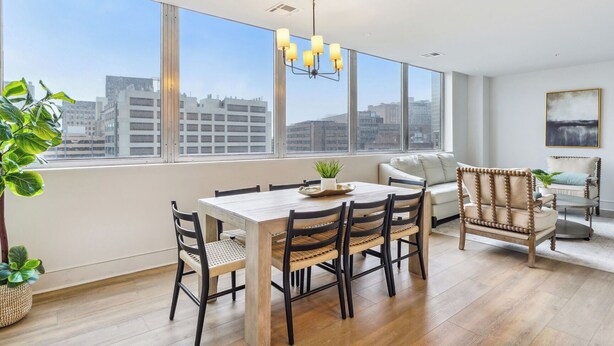 A modern dining area with a wooden table and chairs, a sofa, and a view of the cityscape through large windows.