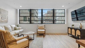 Un salon spacieux avec une grande fenêtre, un canapé, un fauteuil, une table basse et un téléviseur fixé au mur.