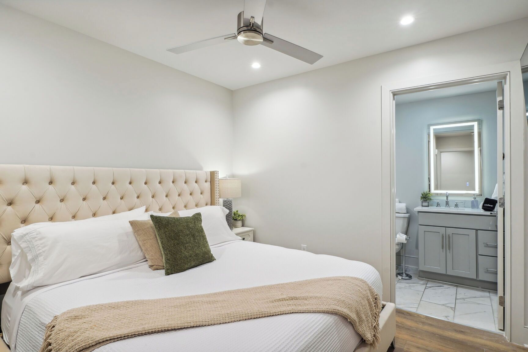 Un dormitorio con cabecera acolchada, cama con ropa de cama blanca, almohada verde, manta color beige, mesita de noche con lámpara, y un baño con espejo, lavamanos y armarios.
