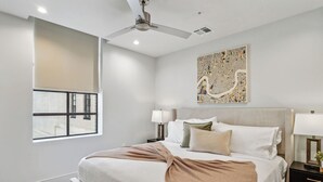 Une chambre d’hôtel moderne dotée d’un grand lit, d’un ventilateur de plafond, d’une œuvre d’art représentant un paysage urbain et d’une fenêtre avec des stores.