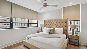 A modern bedroom with a large bed, a ceiling fan, and a view of buildings through the windows. - The Eleanor 10BD/10B, All King Bedrooms w Ensuites (New Orleans)