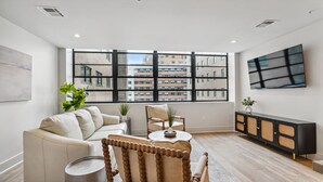 A modern living room with a large window, a sofa, a wooden chair, a small table, and a TV on the wall. - The Eleanor 10BD/10B, All King Bedrooms w Ensuites (New Orleans)