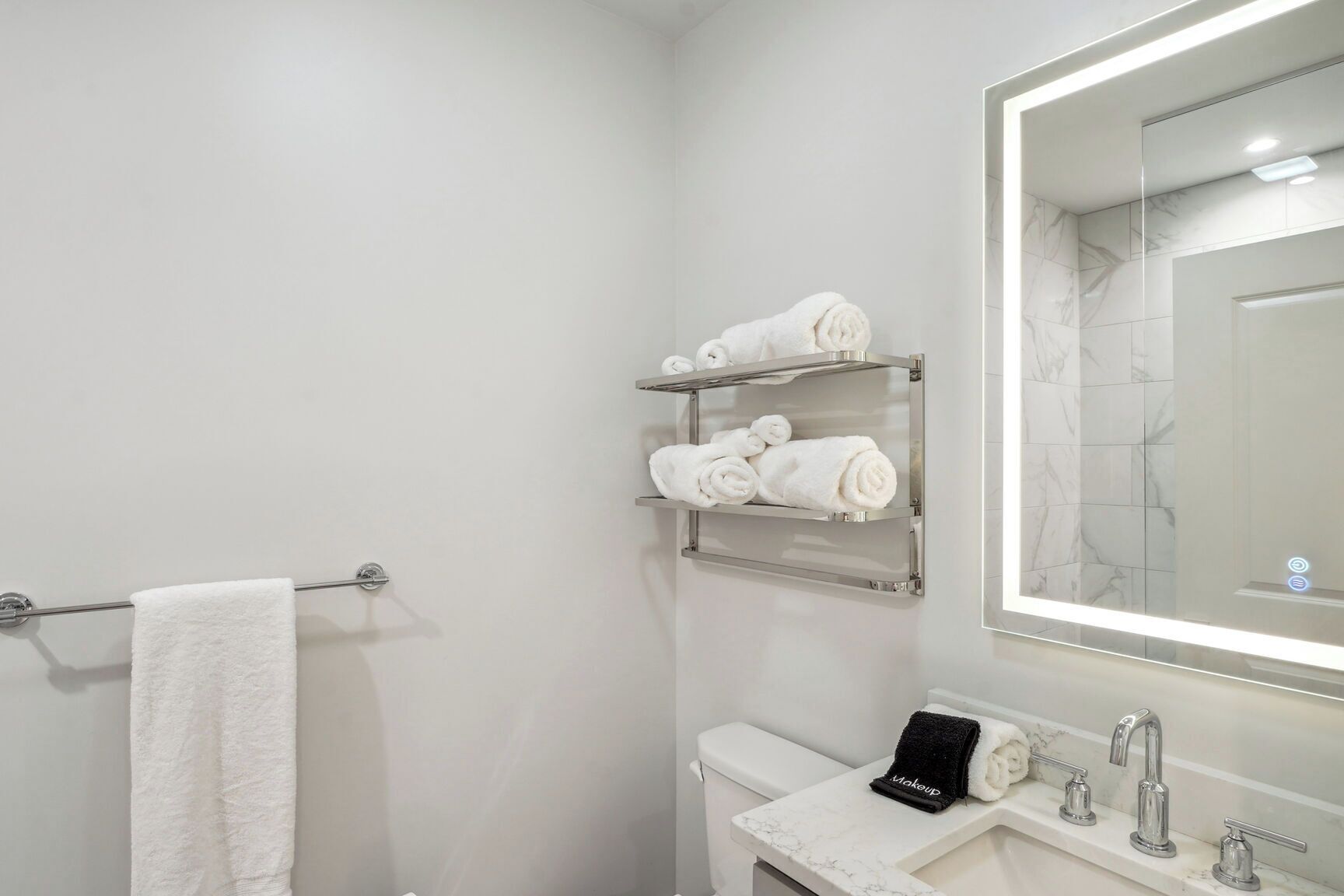 Un baño moderno con inodoro blanco, lavamanos con encimera de mármol, espejo con luz y dos toalleros con toallas dobladas con cuidado.