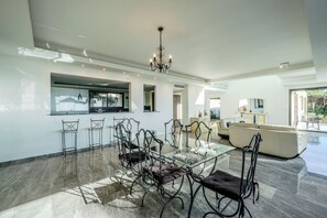 A spacious living area with a glass dining table, modern chairs, a chandelier, and a view of the outdoors.