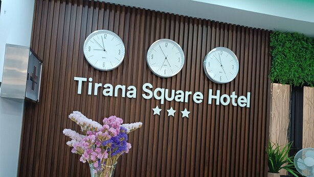 A reception desk at Tirana Square Hotel with three clocks on the wall, a floral arrangement, and a signboard. - Tirana Square Hotel (Tirana)