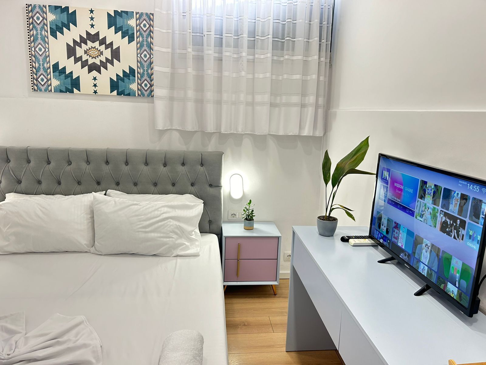 A modern bedroom with a grey tufted headboard bed, a white bedside table with a lamp, a small potted plant, and a TV displaying a social media app.