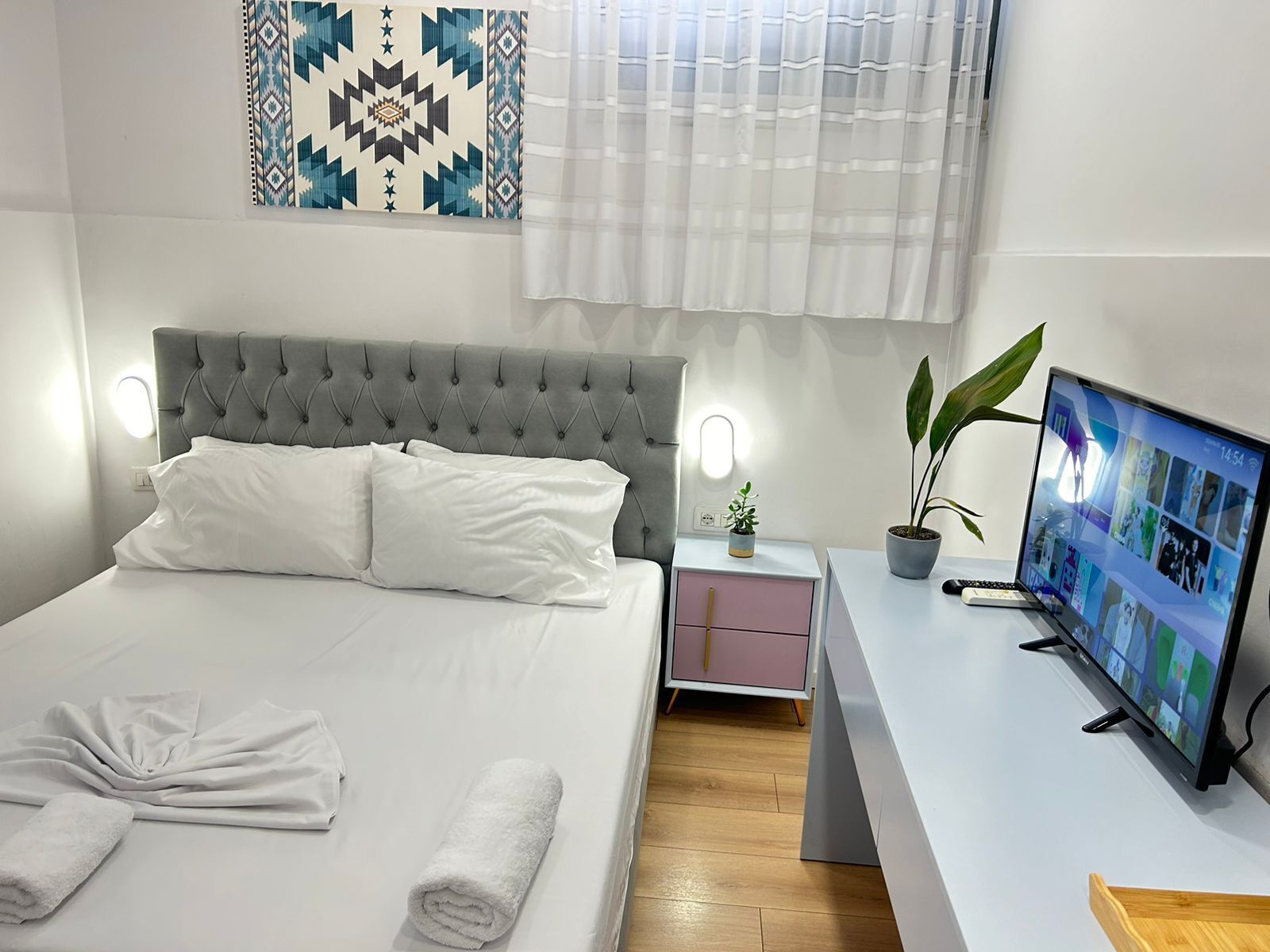 A neatly made bed with pillows and towels, a bedside table with a plant, a TV on a stand, and a patterned wall hanging.