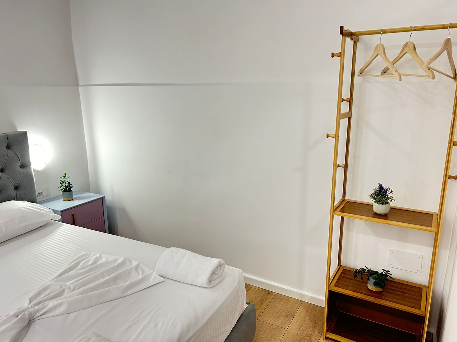 A neatly made bed with white linens, a bedside table with a lamp, and a wooden hanger rack with clothes and potted plants.