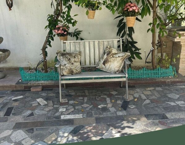 Un banc doté d’une structure métallique et de coussins, entouré d’un sol en pierre à motifs, avec des jardinières suspendues remplies de fleurs et une clôture verte en arrière-plan.