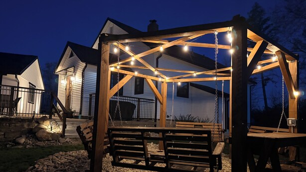 Un pabellón de madera con luces colgantes, un banco y un sendero de piedra.