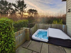 A hot tub in a backyard with a wooden fence, stone tiles, and a view of trees and sky.