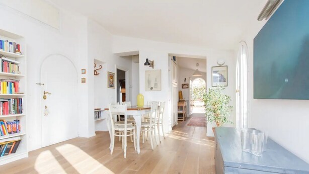 Une pièce avec un plancher en bois, une table à manger blanche avec des chaises, une bibliothèque et une grande fenêtre.