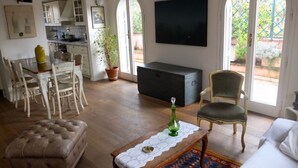 Un salon comprenant un canapé, un pouf, une table et des chaises. Il y a un téléviseur accroché au mur et une plante en pot.