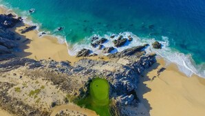 Vue aérienne d’une plage aux eaux turquoise et limpides, bordée d’un rivage sablonneux et d’un terrain de golf verdoyant.