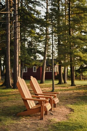 Two wooden Adirondack chairs in a park with trees and a red building in the background.