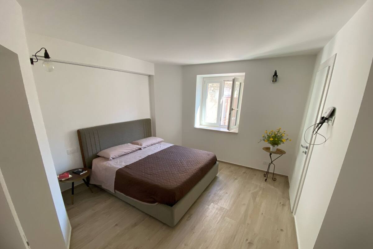 Un dormitorio con cama, mesita de noche, ventana y una pequeña mesa con flores.